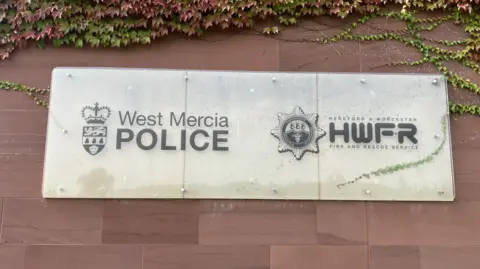 A grey sign fixed to a slate wall reads West Mercia Police and Hereford & Worcester Fire and Rescue Service.