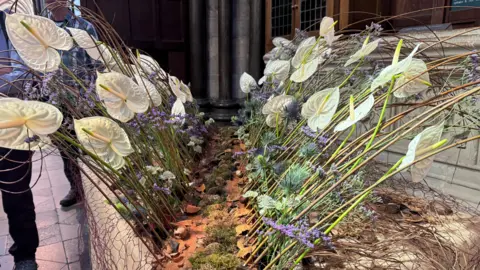 A flower display of white lilies and small purple flowers arranged in what resembles a parting sea. The arrangement is in the main hall in the cathedral.