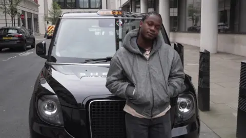 A man wearing a grey hoodie standing in front of a black cab
