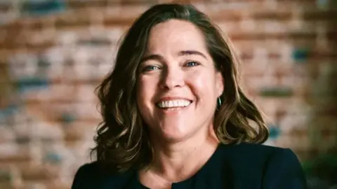 Standards commissioner Dr Melissa McCullough is smiling widely at the camera. She has shoulder-length hair which is brown with caramel highlights. She is wearing a black top. Behind her is an exposed brick wall.