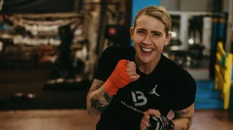 JADE BURDEN Jade, a woman with blonde hair, she wears a black t-shirt and has her hands strapped, she clenches her fist as if she is fighting and smiles brightly.