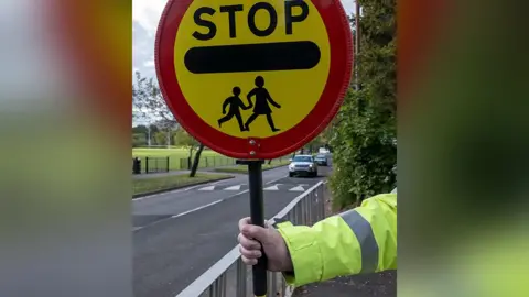 K Neville/Getty Images The arm of an anonymous lollipop warden, wearing a yellow hi-vis coat at a school crossing. The warden is holding a sign telling drivers to stop for children. 