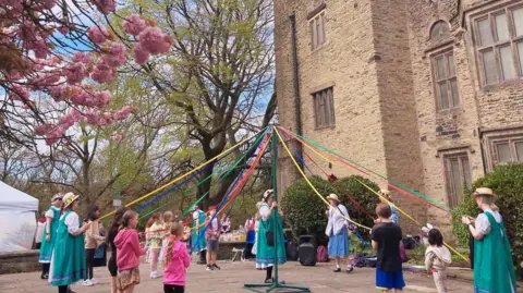 People in traditional green and blue costumes dancing around a maypole outside a large historic stone building, weaving colourful ribbons beneath blooming pink cherry trees on a bright spring day.