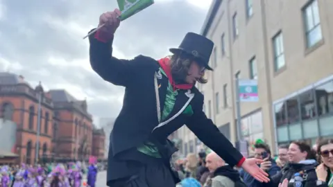 A man on stilts in bending down to shake hands with a child in a crowd. He has a top hat on and wears glasses. Performers in purple are in the background. 