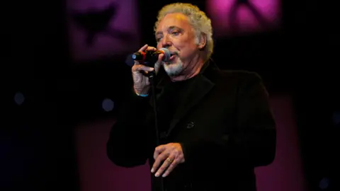 Sir Tom Jones is dressed in all black as he sings into a microphone against a black and pink backdrop. He has curly grey hair.