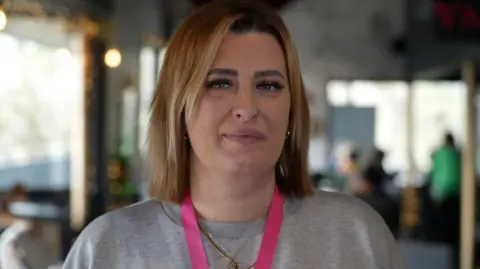 Jamie Niblock Stacey Runciman is standing in the top cafe bar of the New Wolsey Theatre there are windows on both her right and left side. you can also see people at tables in the background. Stacey is wearing a grey jumper with bright pink lanyard around her neck. 