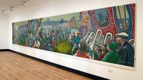 Durham County Council Cornish's mural on a large white wall. It is a long and colourful painting showing people with large banners and musical instruments parading.