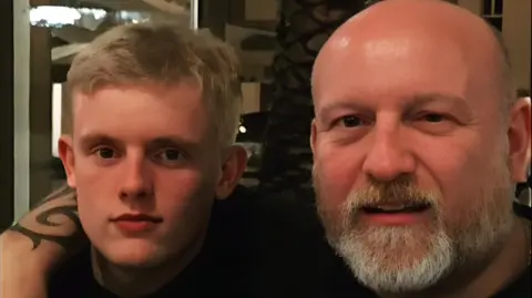 Amber Selvey A father and son next to each in a restaurant, both looking at the camera. The man has a beard and bald head and he has his right arm around a young man who has blonde short hair. 