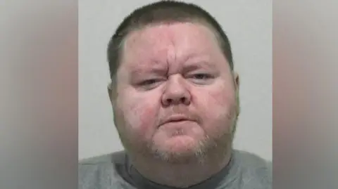 Mugshot of Mark Hogarth. He has a large round head with a stubbly fair beard and short dark hair. he is wearing a grey sweatshirt.