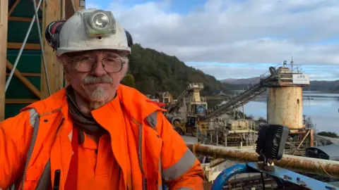 Caroline Thwaites Colin Thwaites is an older man with a neatly trimmed beard and moustache. He is wearing a white hard hat with a lamp fitted to the front, and an orange high visibility jacket. He is outdoors in an industrial area.