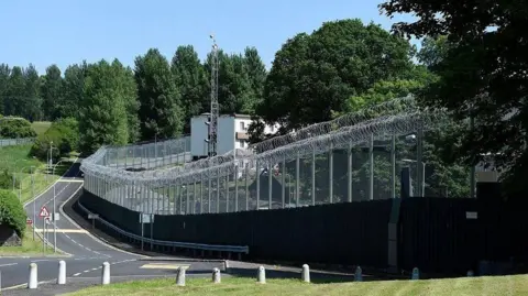 Michael Cooper The perimeter fencing at Hydebank with a road running alongside it with trees throughout the site and a compound behind the wire 