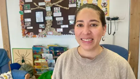 Yingchao wears a beige jumper and smiles at the camera, sitting in front of a children's library area