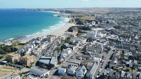 An aerial drone shot of Newquay town centre in the sunshine, with blue skies and the beach on the left, a very blue sea with white waves - and cliffs and countryside stretching into the distance. The buildings are largely grey and white and there are criss-crossing roads visible with cars on them.