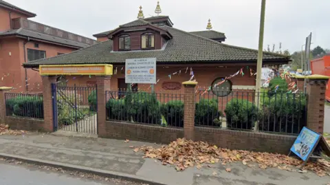 The image shows part of the temple. There is a yellow and orange sign with a metal gate below and adjoining fencing on either side that stands in a low brick wall with trees behind it. The temple has a sloping triangular roof with small windows and and gold-looking ornamental objects in three places on the roof.
