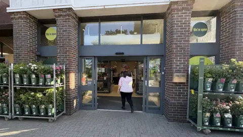 Waitrose A woman walking into a supermarket. There are pot plants placed at the front of the shop. 