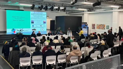 General view of an event. There is a large screen on a black stage and a long table in front of the stage with five people sitting in a row facing the audience. A crowd of people sitting on white chairs are facing the front and a woman in yellow is stood up at the front