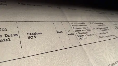 Close-up of an official UK death certificate from the General Register Office, showing typed details and headings, including registration district and cause of death, photographed under soft lighting.
