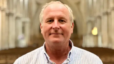 Somerset councillor Mike Rigby. He has short white hair and is pictured inside Wells Cathedral. He is wearing a blue and white striped shirt and is looking at the camera and smiling.