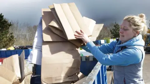 A woman with blonde hair and wearing a light blue waistcoat over a darker blue hoodie, drops pieces of cardboard into a metal bin, painted blue.