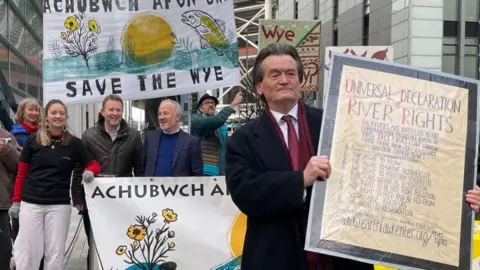 A group of campaigners all holding signs, one of them. One of the signs says Universal Declaration of River Rights, followed by several lines of smaller text, and  another says Save the Wye