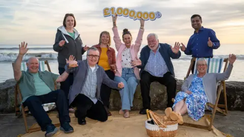 A group of men and women celebrating on a beach with a £1,000,000 sign 