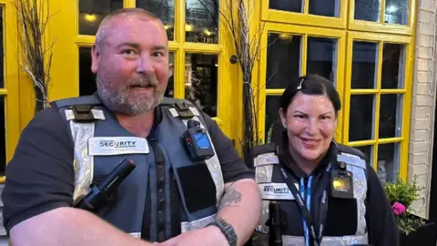 Two security guards with torches and radios in front of a yellow-framed window in Trowbridge