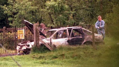 Pacemaker A man in a forensic boiler suit is standing at the rear of a burned out car. The bonet and back door of the car are opened. The vehicle has been destroyed by fire so no longer has any paint, windows or tyres. It is parked in an area surrounded by security and farming fences with lots of grass. There are also trees and a danger sign in the background.