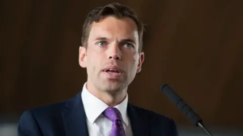 Getty Images Ken Skates in white shirt, navy jacket and purple polka-dot tie, standing behind a mic