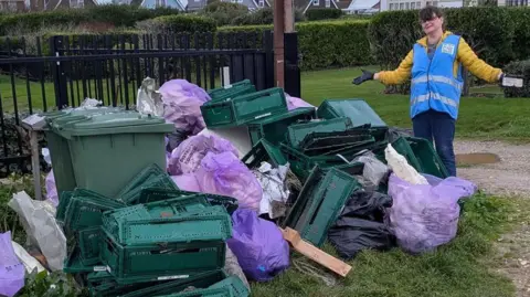 Selsey Beach Litter Ninjas Donna Tretheway stands with arms held wide beside a pile of green plastic crates and purple rubbish sacks on grass.