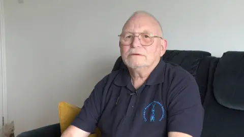 A man wearing a navy polo top is sitting on a sofa and looking toward the camera.