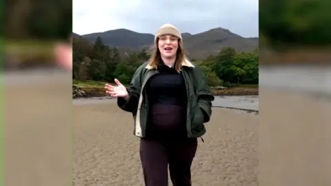 Elle Duffy - pregnant woman in dark clothing, a green jacket and tan hat, walking on a beach under a cloudy sky