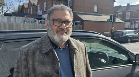 A man with grey hair and beard with dark glasses and a chequed jacket standing in front of a grey car