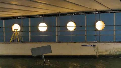 Four circular lights underneath a bridge on Dudbridge Road. The lights are designed to show different phases of the moon and have a warm light.