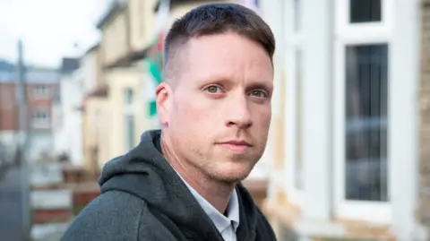 BBC A man with short brown hair and light stubble looks at the camera. Behind him are a row of terraced houses and a Welsh flag. 