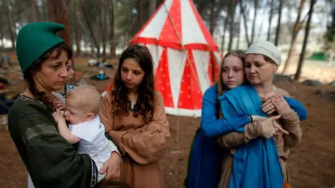 Two women, two girls and a baby dressed in medieval costumers stand in a forest clearing in front of a red and white, circular tent. The woman on the left wears a green smock and cap and is holding a baby dressed in white. A girl wearing a brown dress with long brown hair is watching her. To the right, a girl dressed in blue, with long blond hair, hugs a woman wearing a brown dress and white cap.