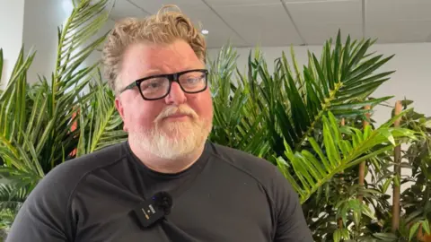 A man with a bear and wearing black-rimmed glasses stands in front of some green houseplants inside while being interviewed 