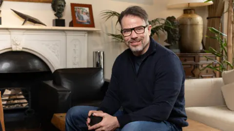 Nick Saffell/University of Cambridge A middle-aged man wearing glasses, a dark blue zip-up hoodie and blue jeans is sat on a coffee table in a living room. He's holding a phone in his hands and smiling at the camera. 