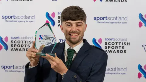 Fin Graham - a young man with brown hair - smiling at camera and holding up a glass award trophy. He is standing in front of a 'Sportscotland Scottish Sports Awards' banner.
