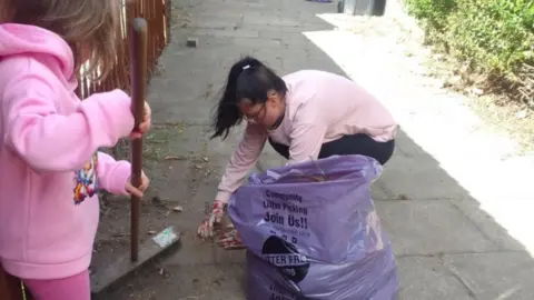 Laura Brennan An image of a woman in a pink jumper and black leggings, picking up rubbish next to a purple bin bag with a young girl in pink, holding a broom, next to her. 