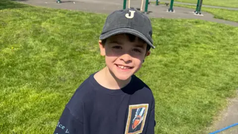 NHS Greater Glasgow and Clyde/ PA A small boy with a blue baseball cap. He is in a park on a sunny day, smiling at the camera