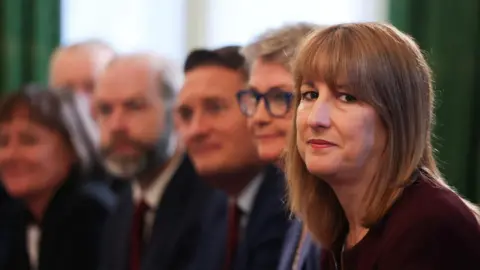 PA Media Rachel sits on the right of the frame, wearing a dark maroon top, looking seriously into the camera. Behind her sit several cabinet ministers, who appear blurry.