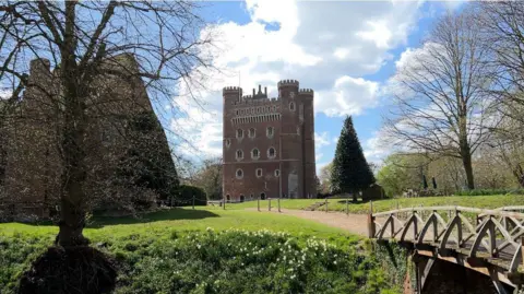 BBC News A tall castle sits in the centre of the picture, surrounded by trees, grass and a bridge on a sunny day with clouds behind it