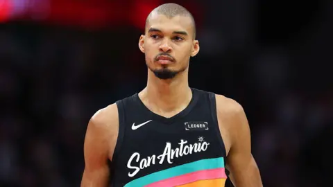 Victor Wembanyama wearing a vest that is black with blue, pink and orange stripes and the words San Antonio across the front