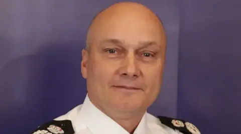 Headshot of Nick Dean in his uniform. He is against a blue background and is smiling at the camera. 
