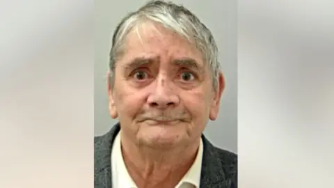 Grey-haired Brian Trevithick, wearing a grey suit and white shirt, purses his lip as he looks straight ahead in his police mugshot