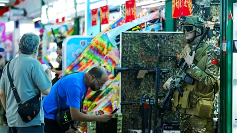 Xiqing Wang, Xiqing Wang Two men look at a stall that has a life-size toy soldier standing guard, with a toy gun.