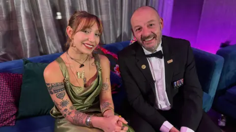 Two people are seated on a plush blue sofa in a lounge setting with purple ambient lighting. The woman is wearing a shiny green satin gown with thin straps and layered necklaces, including a dolphin pendant. The man is dressed in a formal black tuxedo with a white shirt and bow tie, and several medal-style decorations are pinned to the left side of the jacket. Behind them is a backdrop of silver-grey curtains, and colorful cushions in red and green add contrast to the seating area. Both are smiling.
