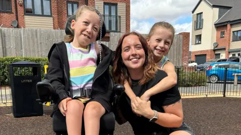 A young Mum with her two daughters in an enclosed park. One is in a wheelchair on the left while the other is hugging the Mum on the right