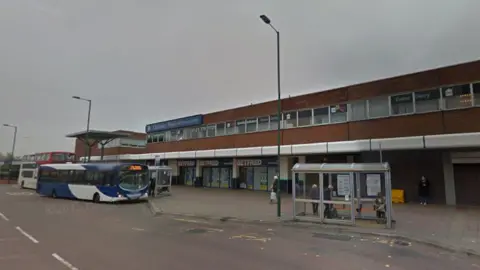 A blue and white single-decker bus is stopped at a bus station which has a row of shops behind. There are people standing at a bus shelter on the right hand side of the image. Three tall lampposts can be seen. The sky is grey and cloudy.