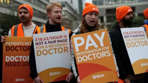 A line of people wearing orange hats and carrying placards saying Pay Restoration for Doctors and £18.62/Hour is not a fair wage for a resident doctor